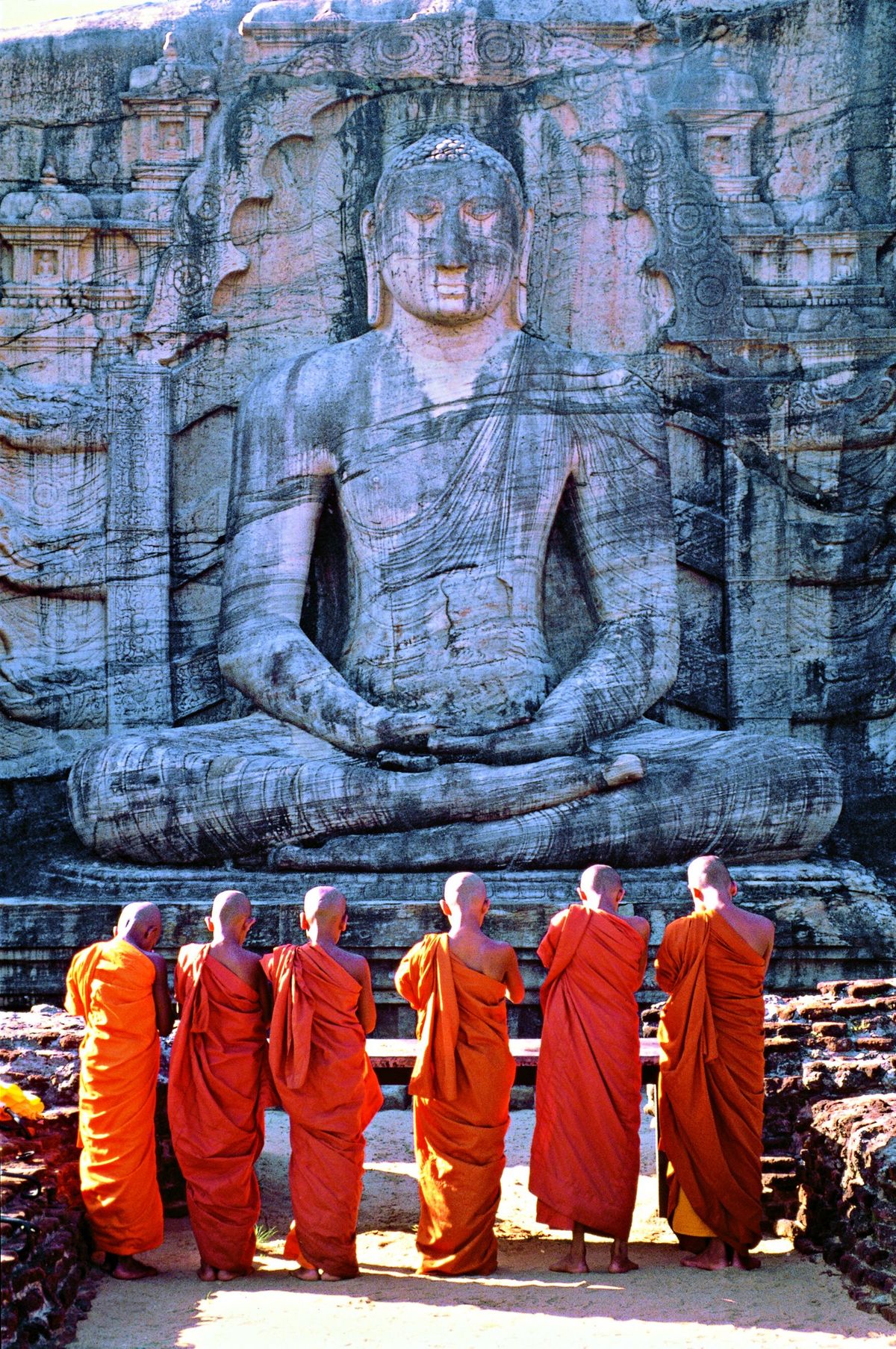 Six monks in orange robes stand before the seated Buddha carved into the rock at Gal Vihara, Polonnaruwa