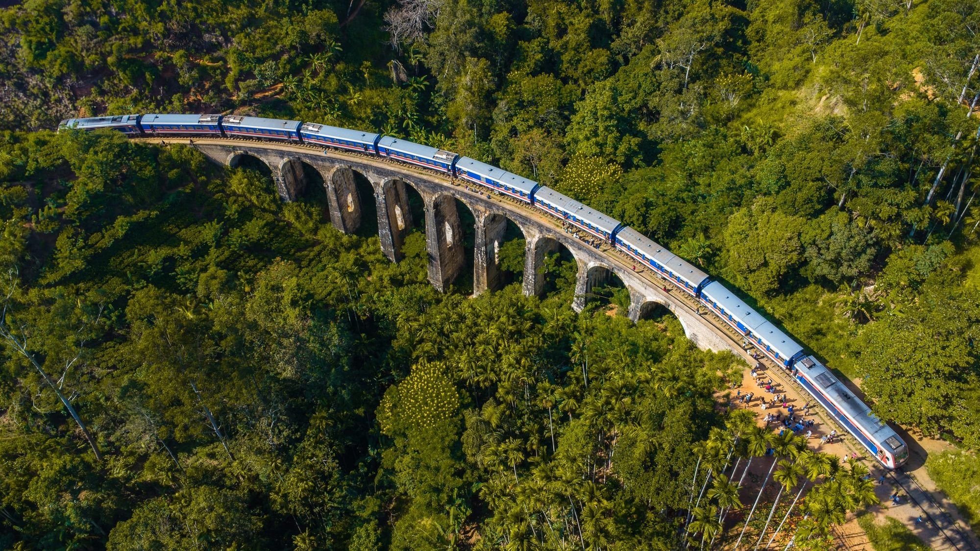 A blue train crossing the Nine Arch Bridge through thick forest, from the air