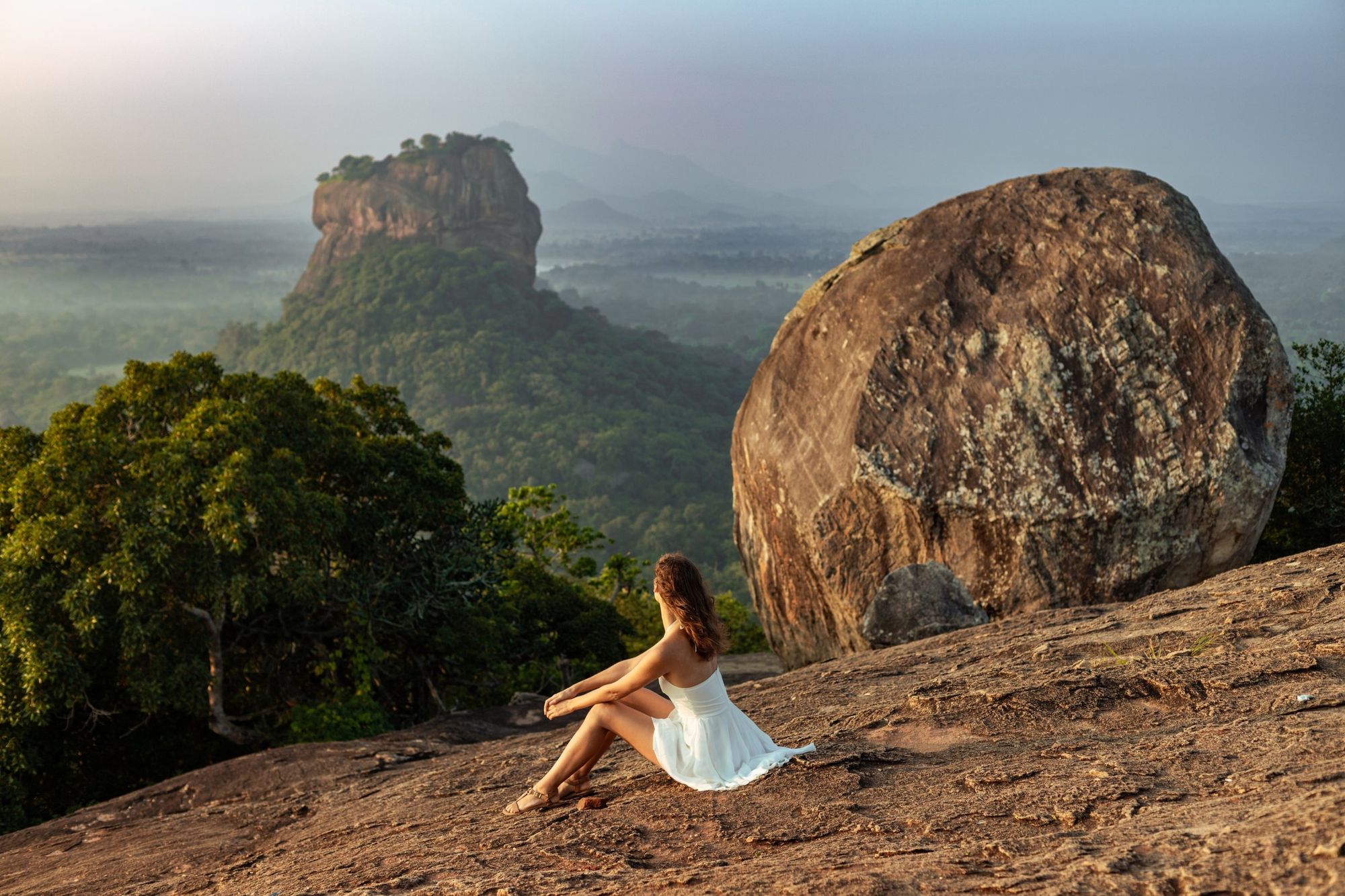A woman sits on the rock at the top of Pidurangala, looking across to Sigiriya at sunrise