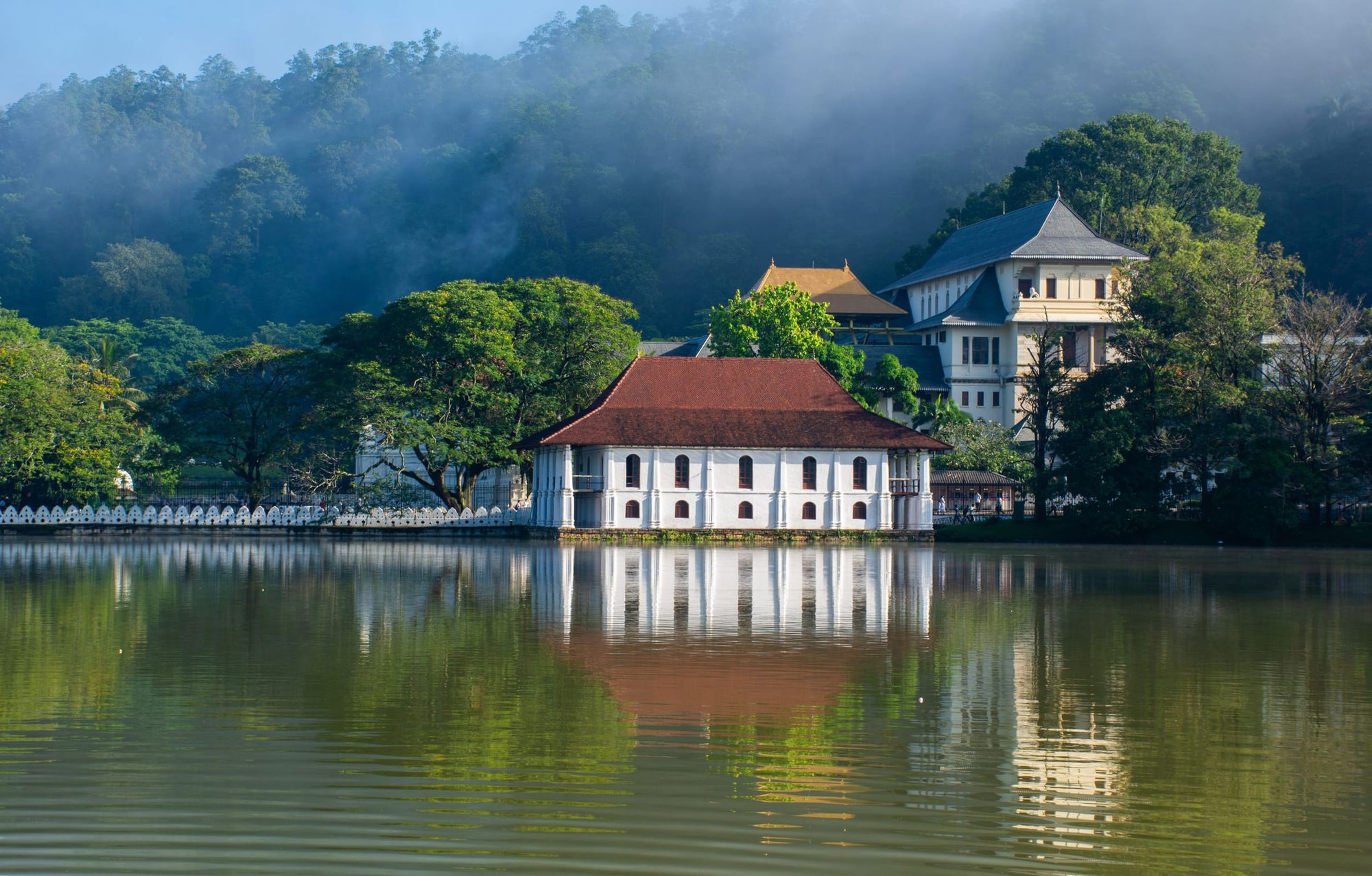 The Temple of the Tooth on the shore of Kandy lake, reflected in still water, hills in mist behind